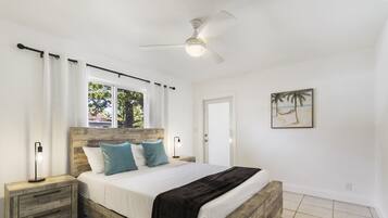 A bedroom with a wooden bed frame, white bedding, a ceiling fan, and a beach-themed painting on the wall.