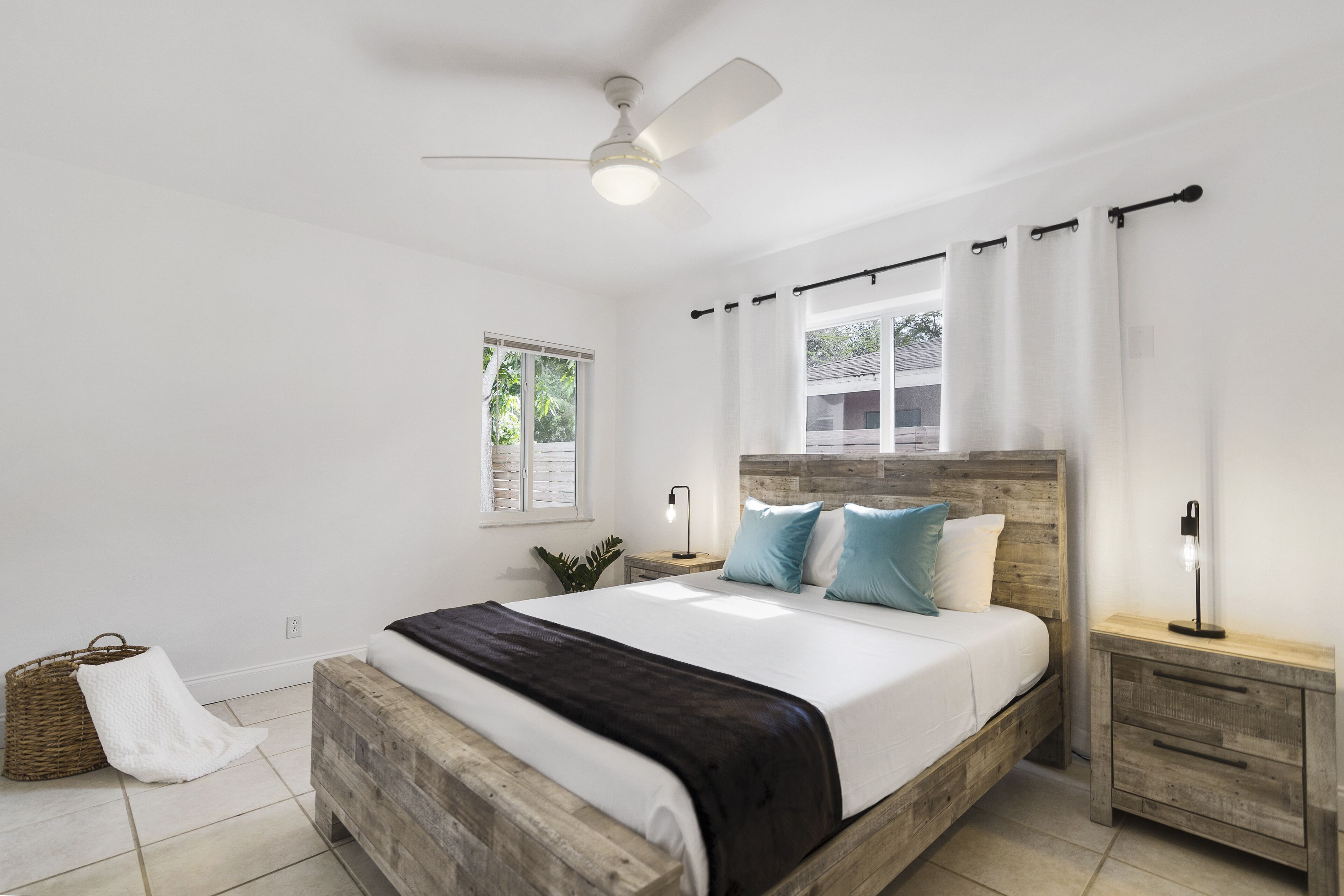 A bedroom with a wooden bed, a ceiling fan, a nightstand with a lamp, and a window with curtains.
