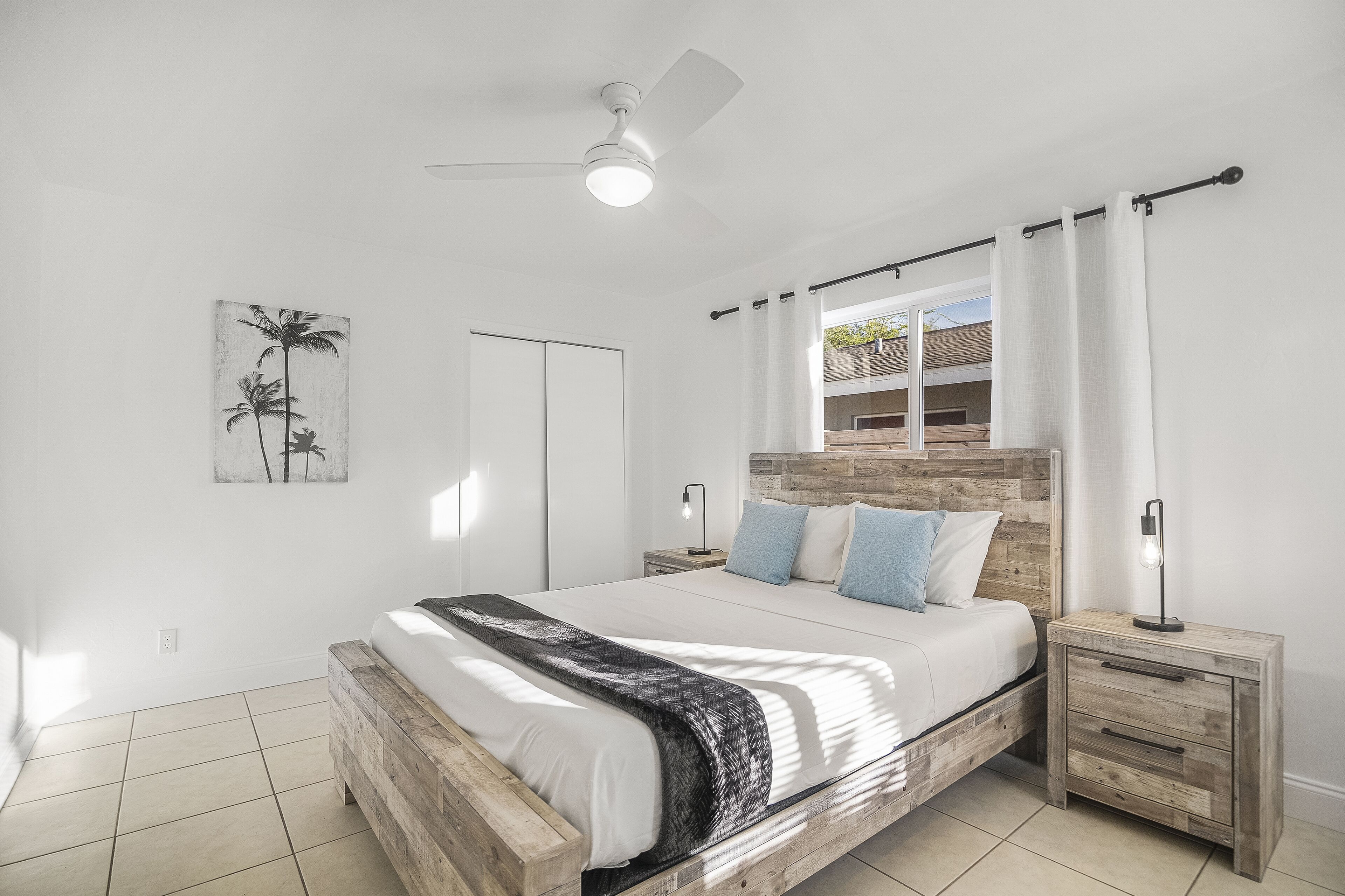 A bedroom with a wooden bed frame, white bedding, a ceiling fan, and a nightstand with a lamp.