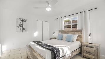 A bedroom with a wooden bed frame, white bedding, a ceiling fan, and a nightstand with a lamp.