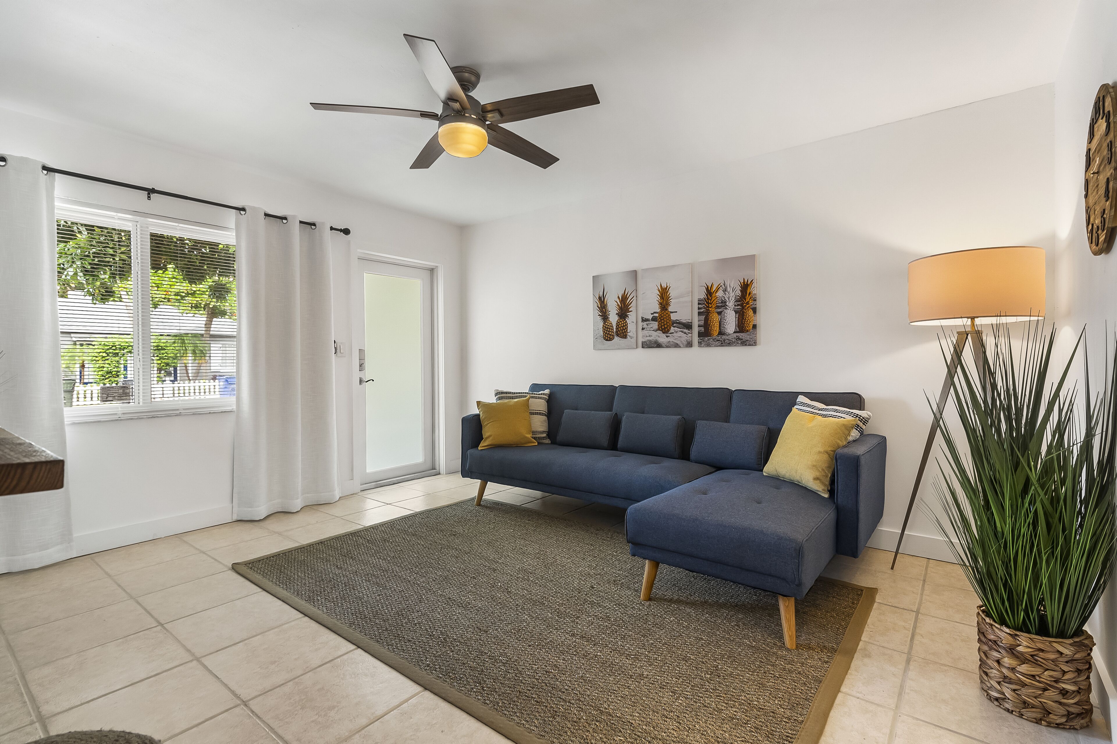 A living room with a dark blue sectional sofa, a ceiling fan, a large potted plant, and a wall clock.