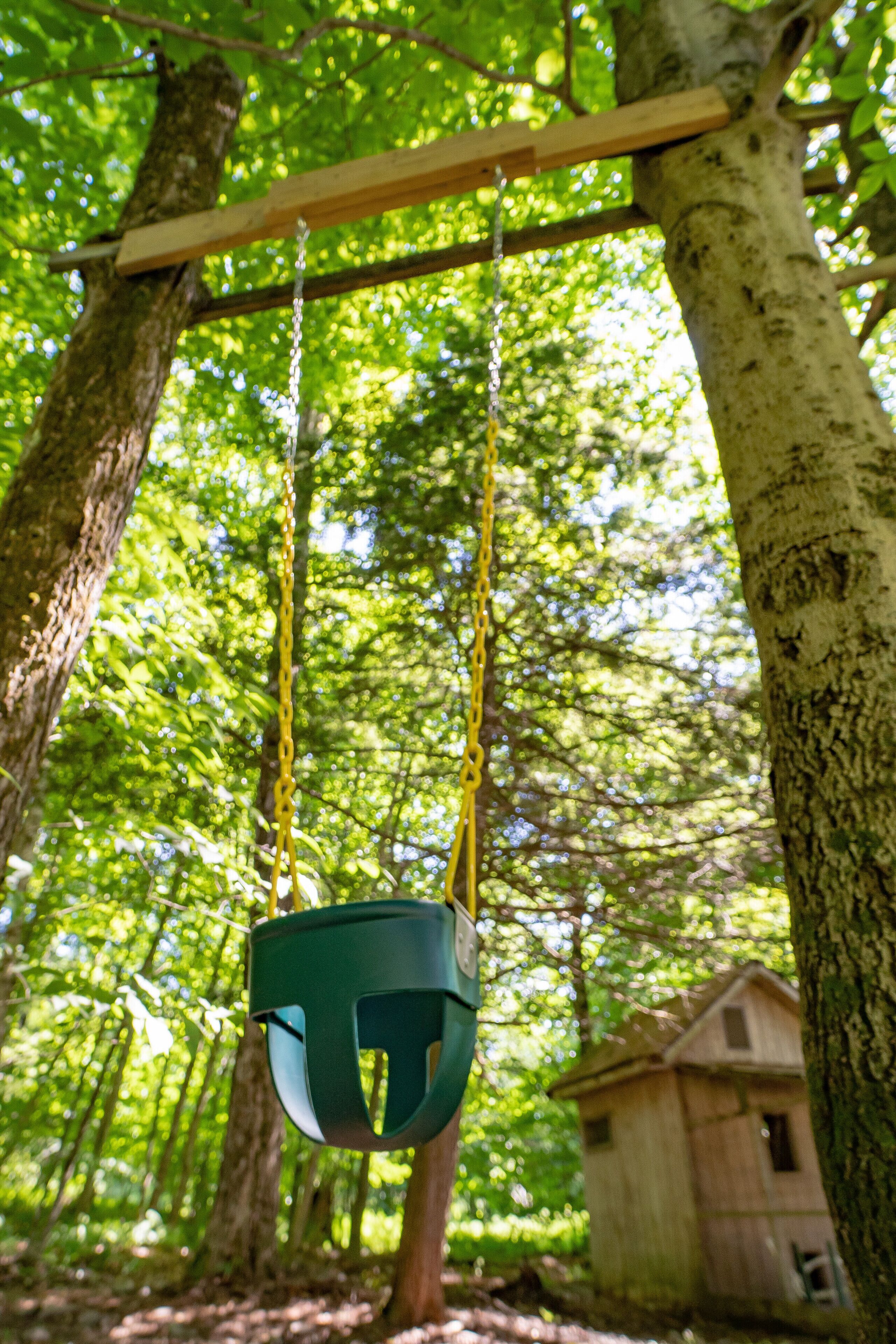 A wooden swing suspended by yellow ropes in a forested area with a wooden structure in the background.