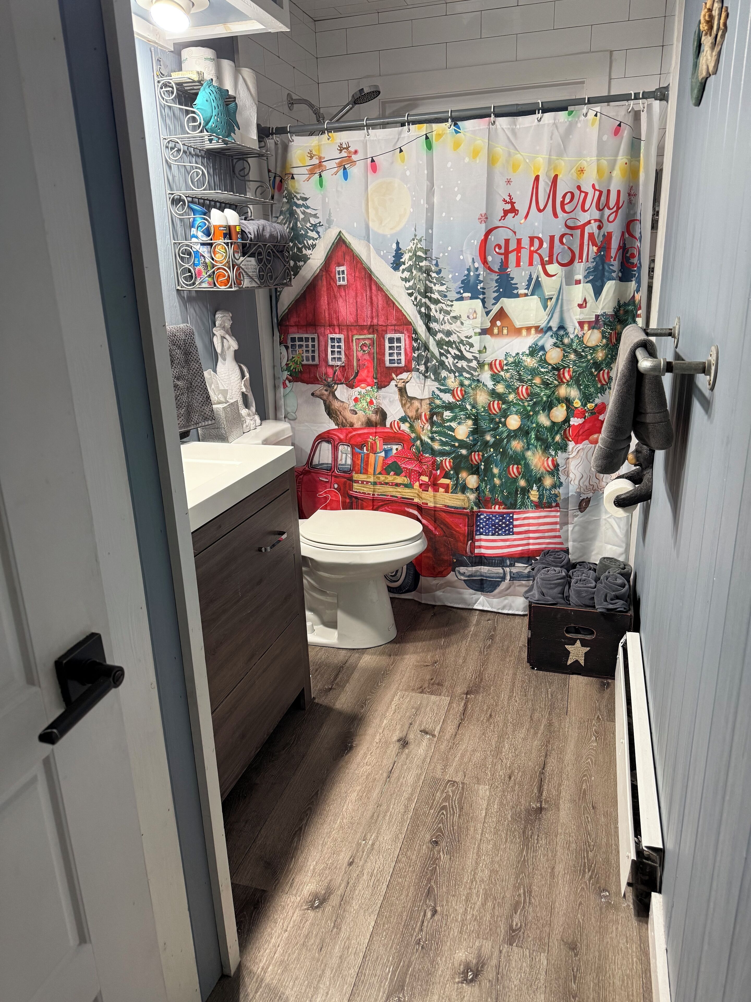 Een badkamer met een houten vloer, een wit toilet, een rode woondecoratie, een kerstboom, en een douchegordijn met de tekst 'Merry Christmas'.