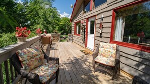 Eine Holzterrasse mit Korbstühlen und einem Tisch, mit Blick auf einen üppigen Garten und ein Haus mit roter Tür und Fenstern.