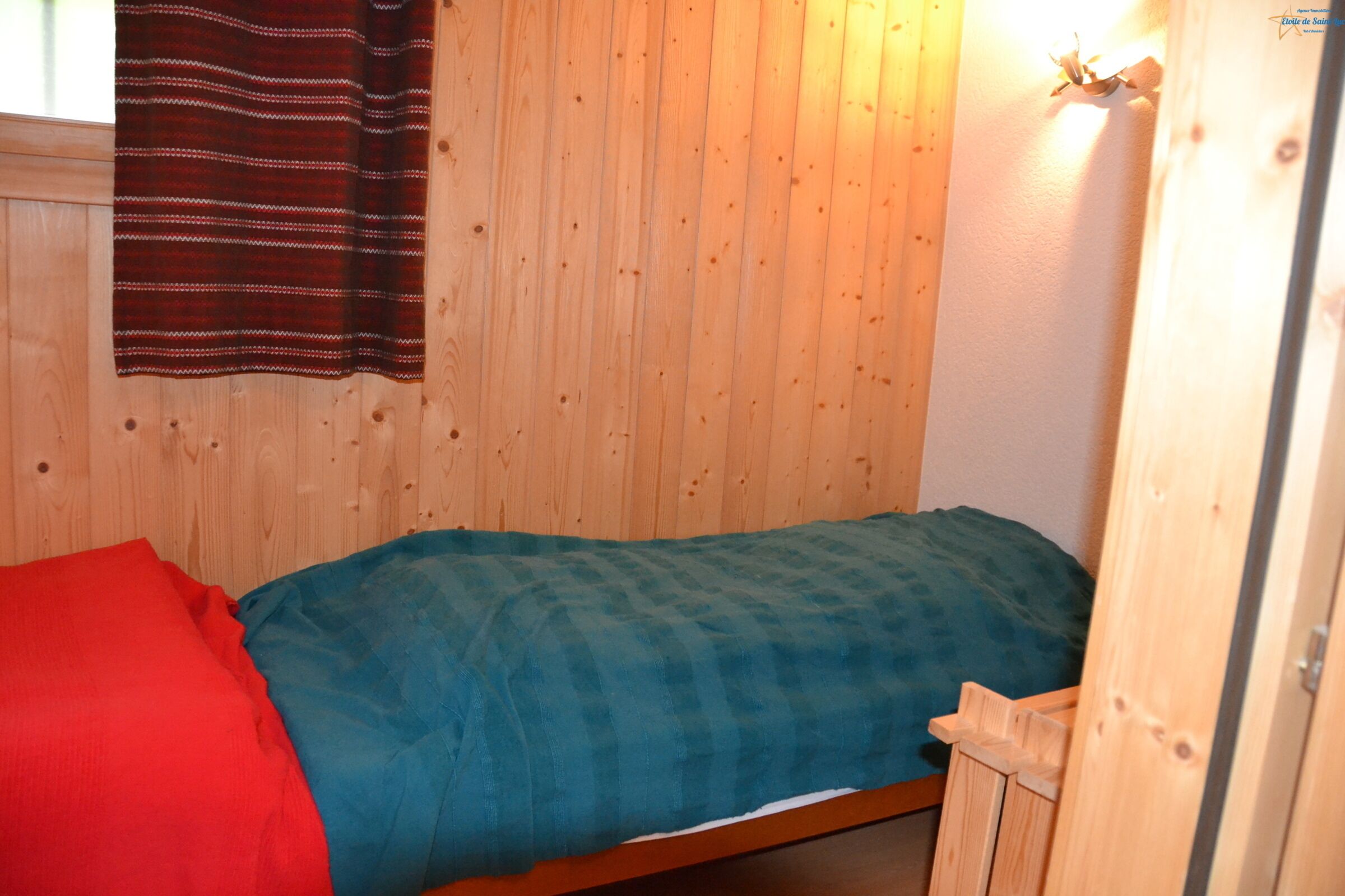 A single bed with a blue and red blanket, wooden walls, and a window with a striped curtain.