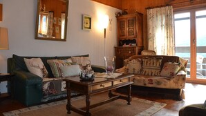 A cozy living room with a green sofa, a wooden coffee table, and a patterned armchair. A large mirror and a cabinet with decorative items are also visible. - Outside : Apartment for 4-6 people, balcony with beautiful view -Inside : 65 sqm, living with , dine (St-Luc)