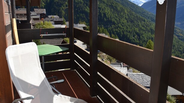A balcony with a wooden railing, a white chair, and a view of a mountainous landscape. - Outside : Apartment for 4-6 people, balcony with beautiful view -Inside : 65 sqm, living with , dine (St-Luc)