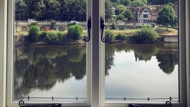 A window with a view of a calm lake, trees, and a traditional building across the water. - The Courtyard (Bewdley)
