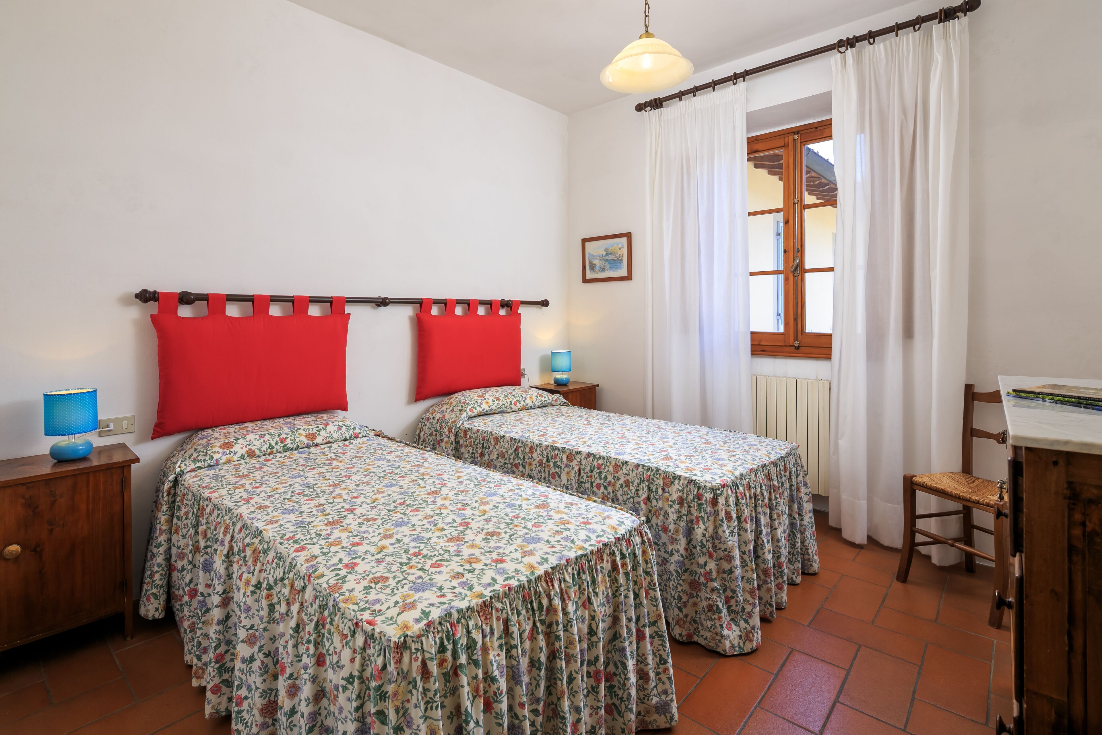 Ein Zimmer mit zwei Betten, einem Holztisch, einem Stuhl, einer Lampe und einem fenster mit Vorhängen.