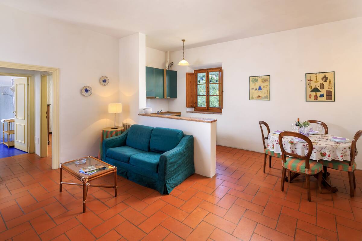 Un espace de vie compact comprenant un canapé, une petite table, un coin repas avec des chaises, un coin cuisine avec des placards et une fenêtre donnant sur un paysage agréable.