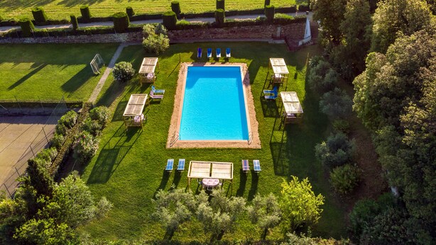 Vue aérienne d’un espace piscine avec une piscine rectangulaire, entourée de pelouse et d’arbres soigneusement entretenus. On y voit plusieurs chaises longues et une petite table sur laquelle est posé un drapeau.