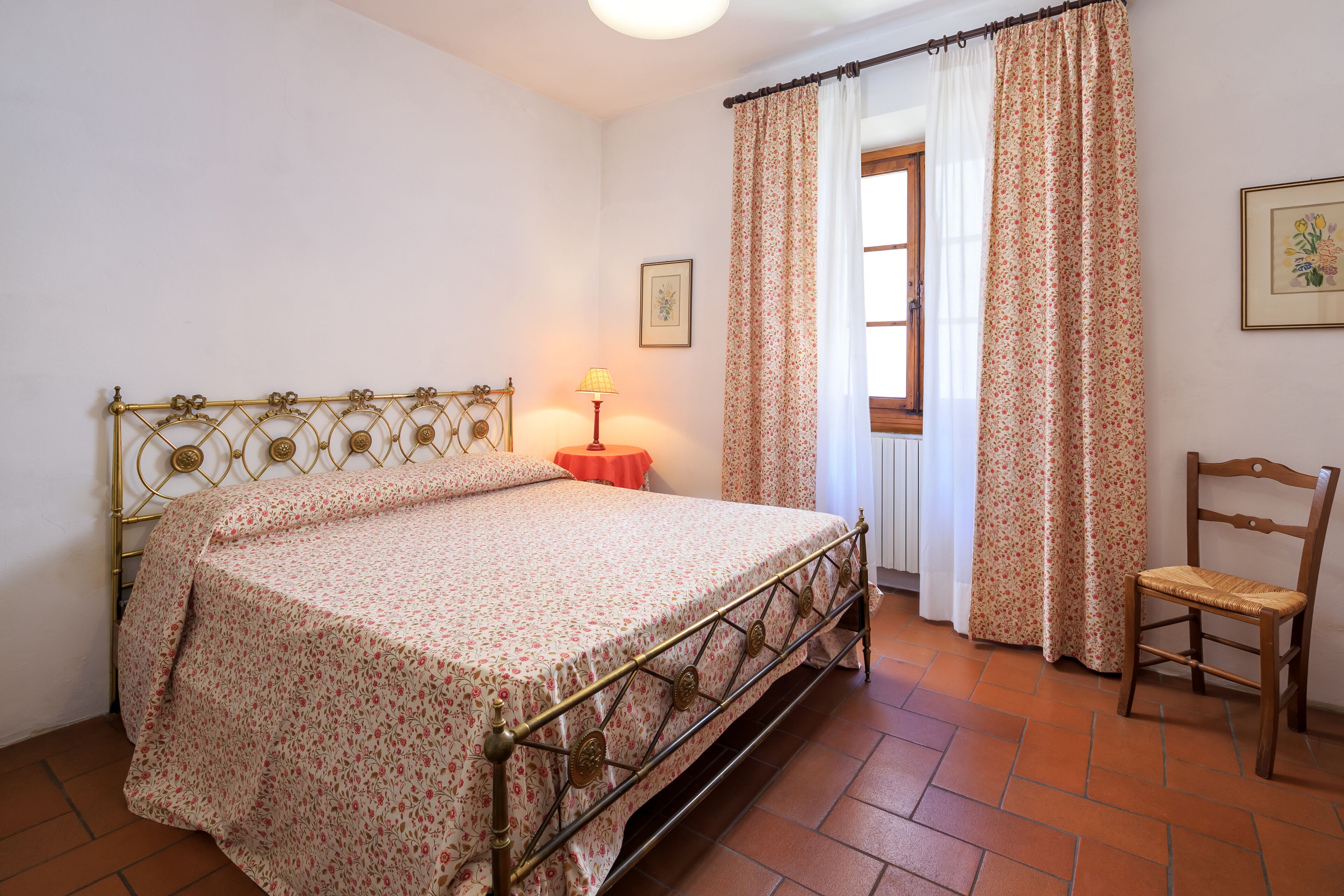 Un dormitorio con cama, una silla, una lámpara y una ventana con cortinas.