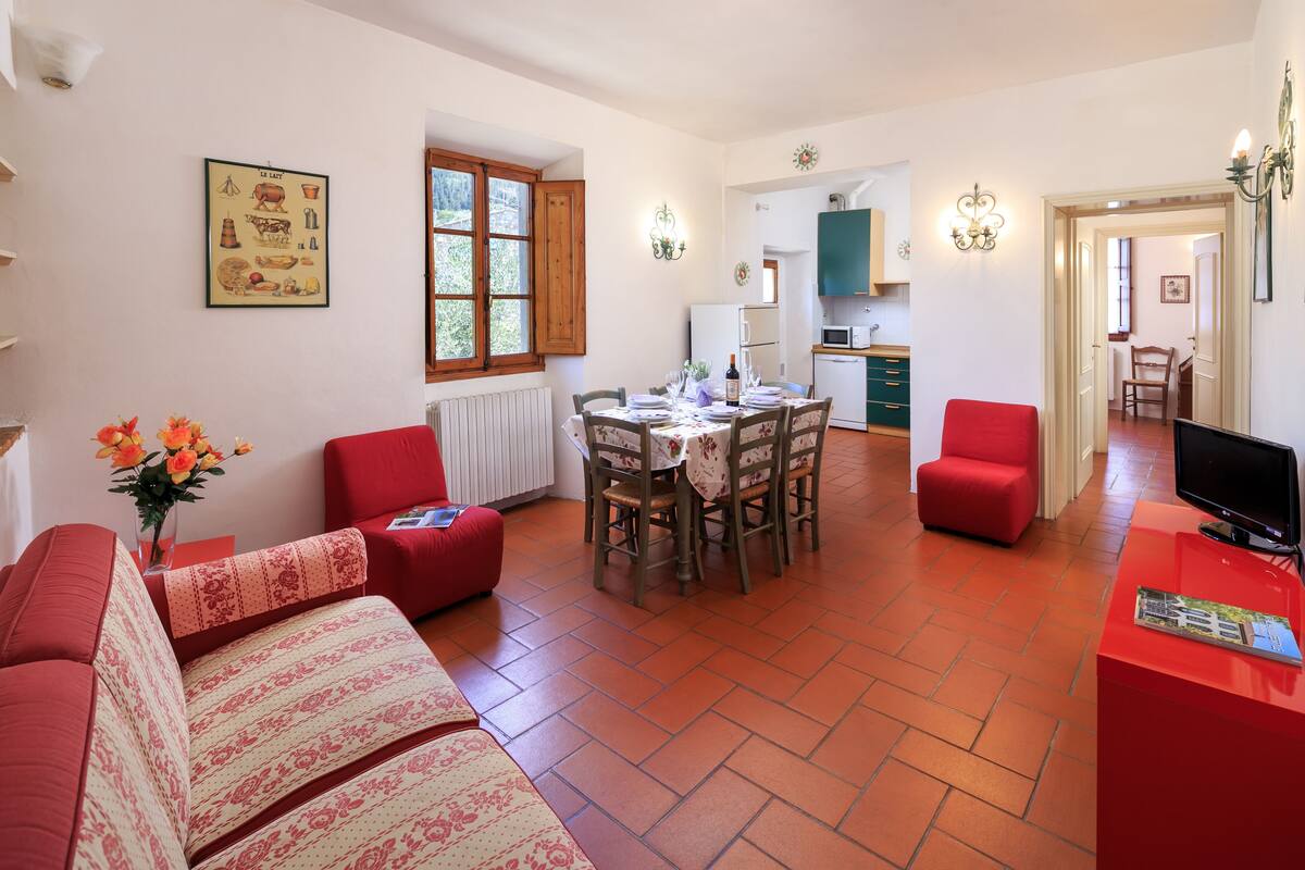 Un salon chaleureux avec un canapé rouge, une table à manger avec des chaises et un téléviseur posé sur un meuble rouge.