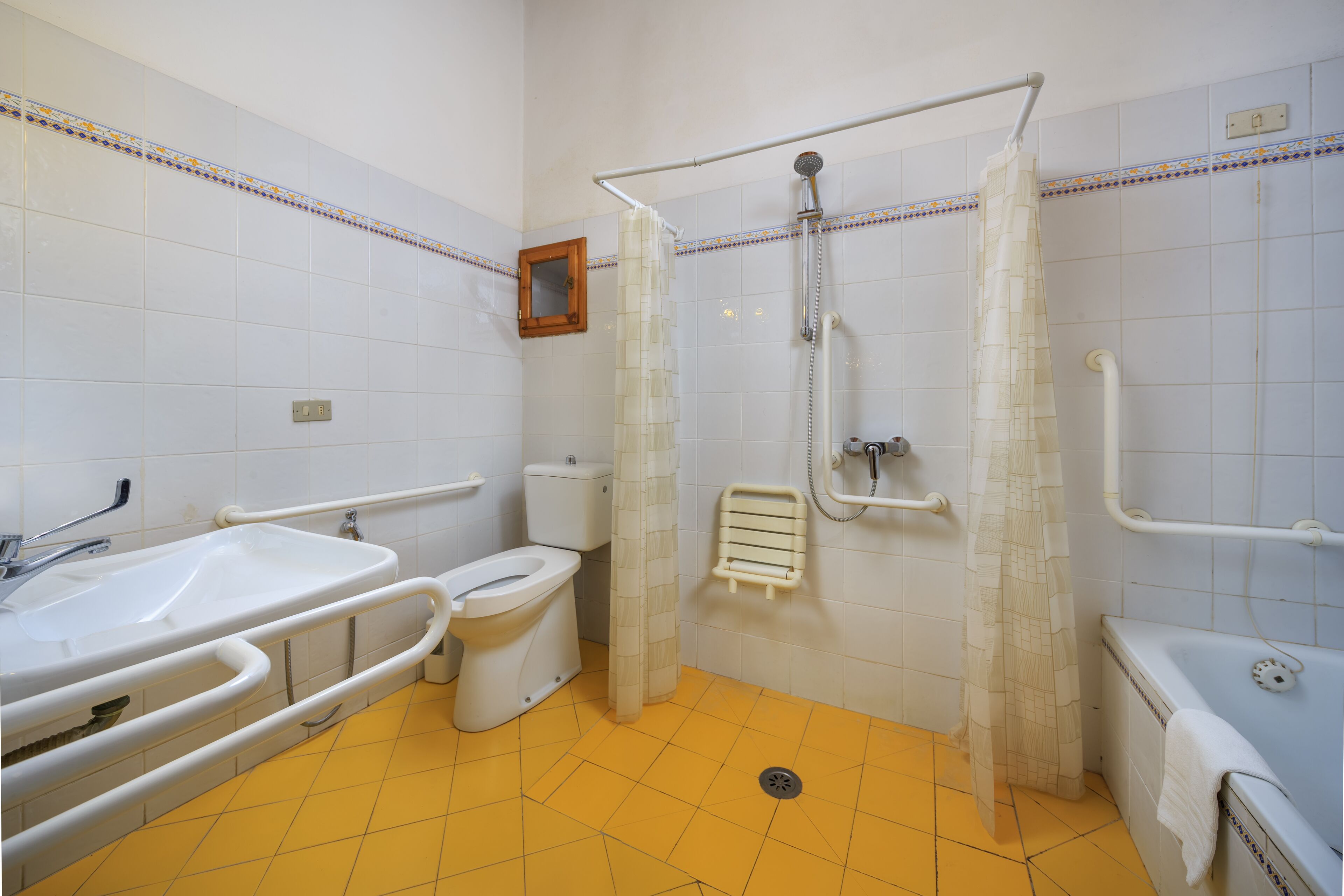 A bathroom with a yellow floor, white tiles, a sink, toilet, shower, and grab bars.