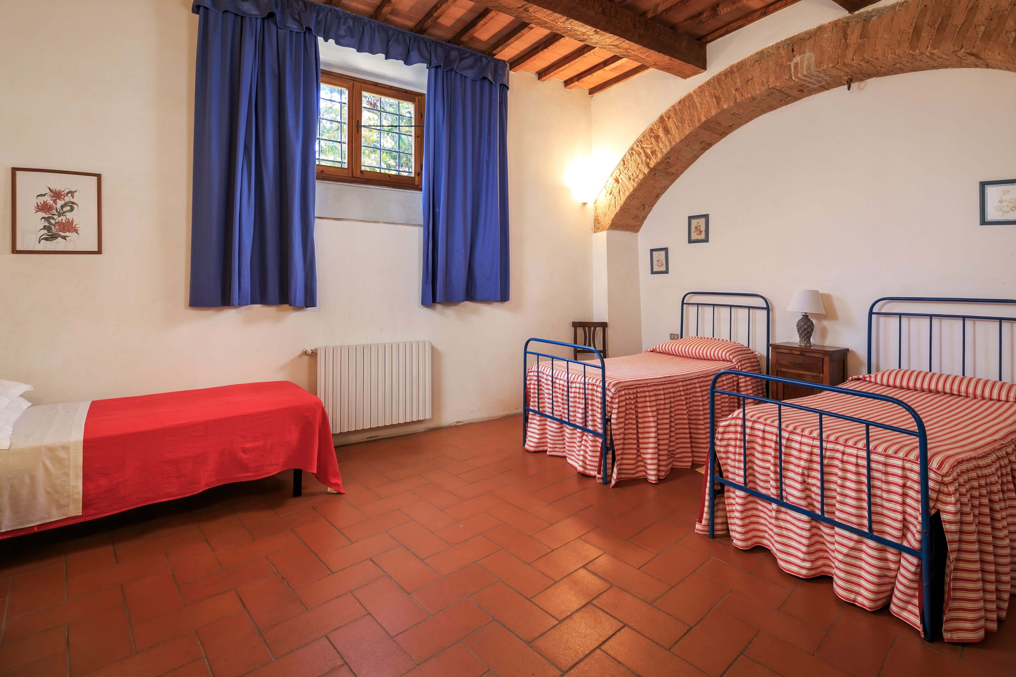 A room with two single beds, a window with blue curtains, a radiator, and a brick floor.