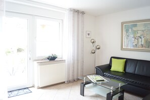 A modern living room with a glass coffee table, a black leather sofa with a green pillow, a painting on the wall, and a window with curtains.