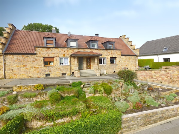 A stone house with a tiled roof, a front porch, and a garden with various plants.