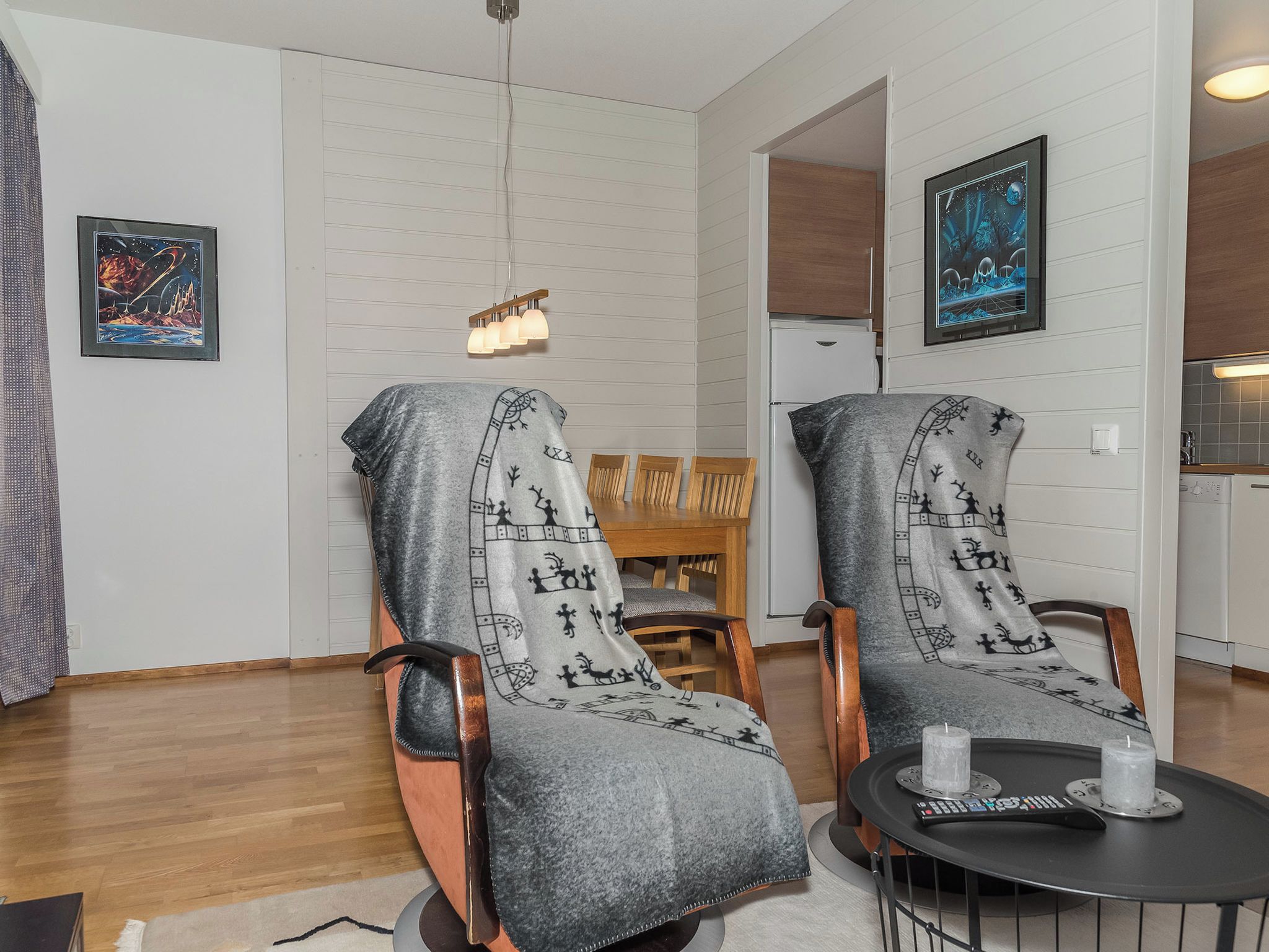 Uma sala de estar moderna com dois poltronas cobertas por um cobertor, uma mesa de jantar com cadeiras, uma pequena mesa redonda com um controle remoto e obras de arte nas paredes.