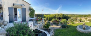 Una casa de piedra con puerta gris y un pequeño jardín con estanque y diversas plantas.