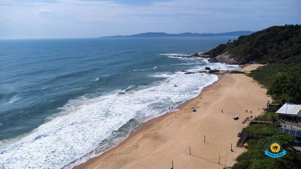 A beach with a sandy shore, a few people, and a building with a logo on the right. - Paraíso do Estaleirinho - Baln Camboriú (Balneário Camboriú)