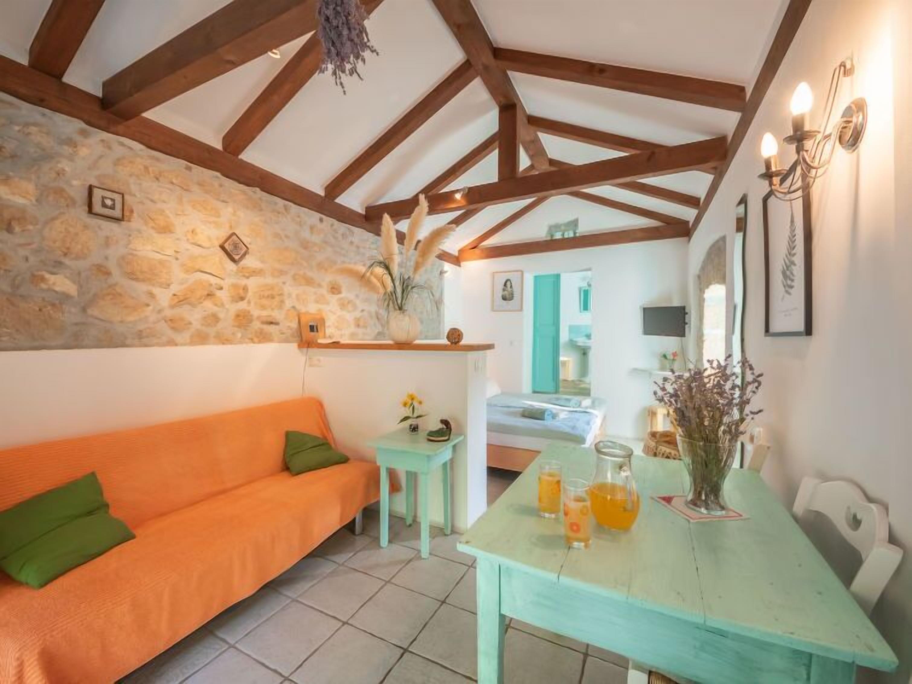 Une pièce chaleureuse avec un mur en pierre, des poutres apparentes, un canapé orange, une petite table avec des boissons et une vue sur une autre pièce.