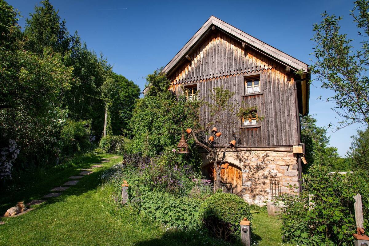 Ein rustikales Holzhaus mit Stein Fundament, umgeben von üppiger Vegetation und einem Kiesweg.