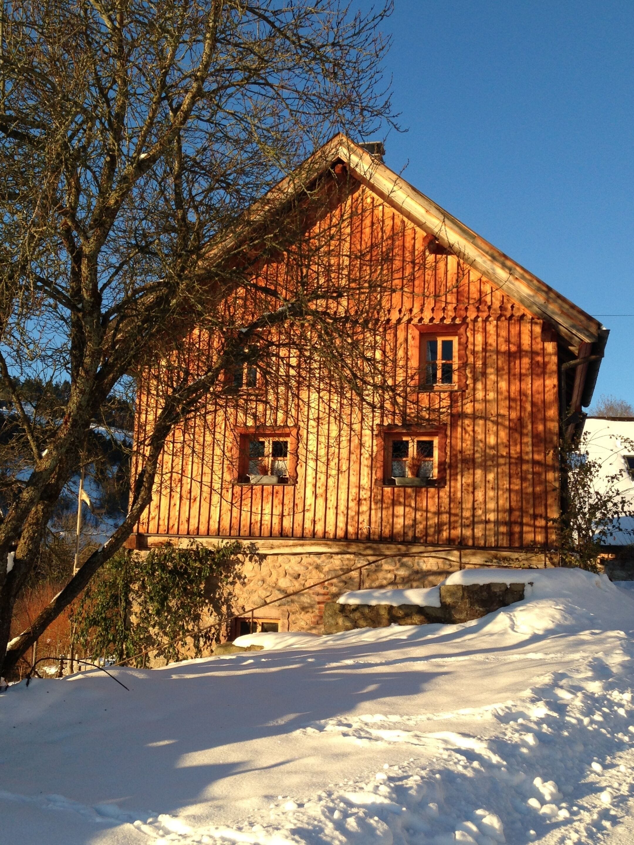 A wooden cabin with a stone foundation, surrounded by snow and trees.