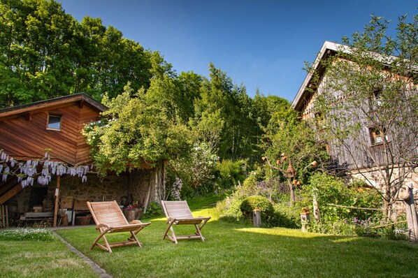 A rustic wooden house with a stone foundation, two wooden lounge chairs, and a lush garden with various plants and trees. - La Grange D'hannah - Gîte & Chambre D'hôtes de Charme (Orbey)