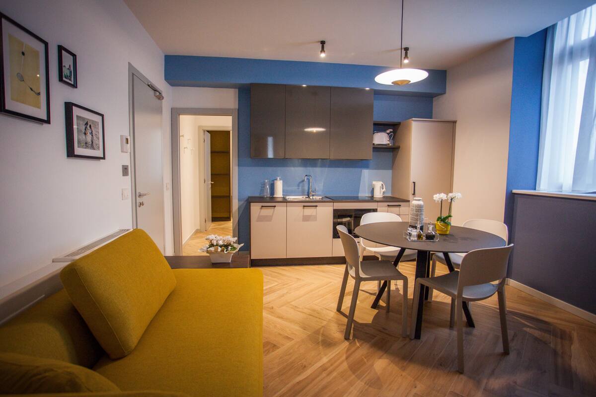 Een moderne woonkamer met een geel bankje, een eethoek met een tafel en stoelen, een keuken met donkere kastjes en een groot raam met gordijnen.