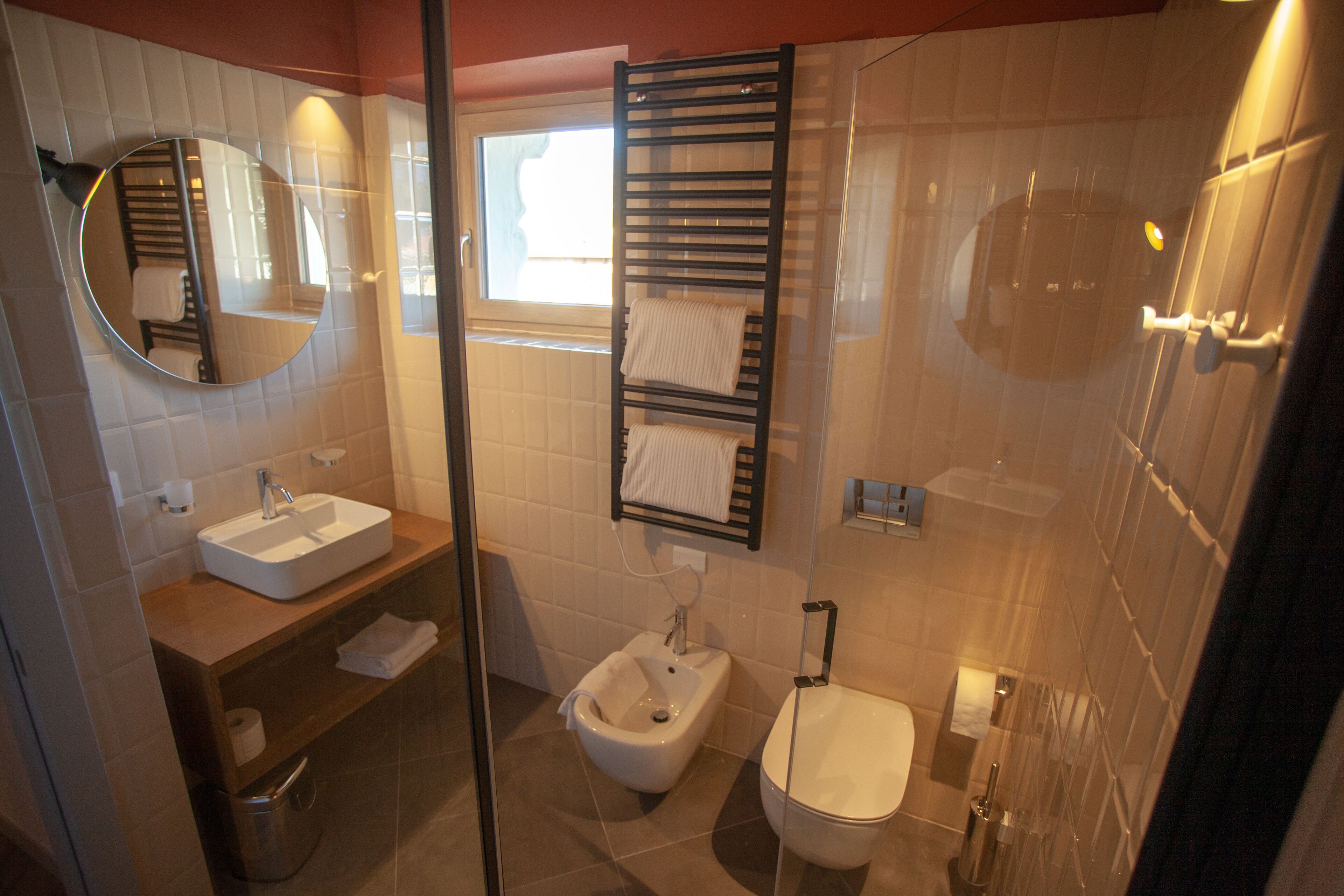 Une salle de bain moderne équipée d’un lavabo, d’un bidet et de toilettes. On y trouve un porte-serviettes avec des serviettes, un miroir et une fenêtre qui laisse entrer la lumière naturelle.