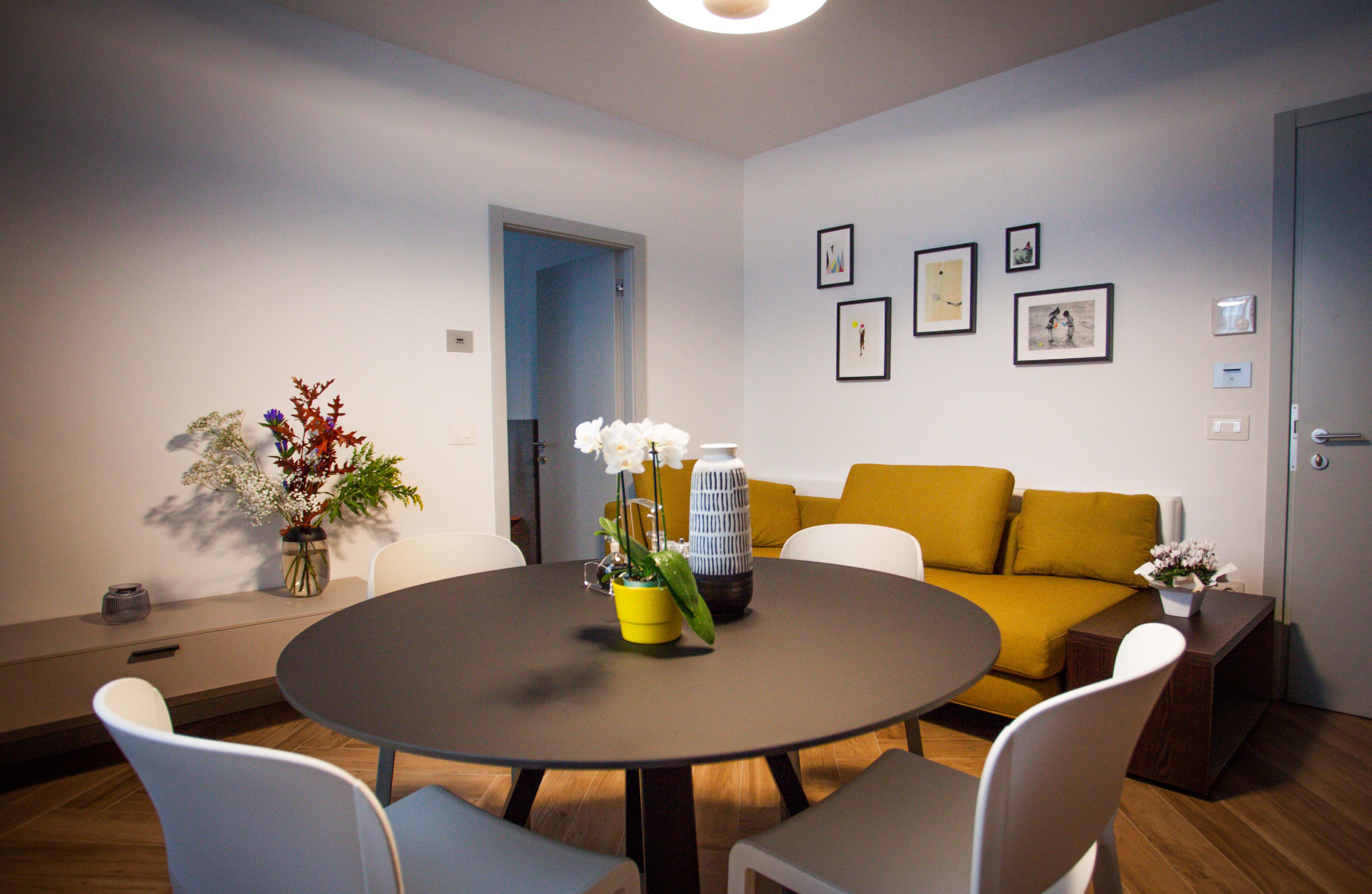 A modern living room with a round table, white chairs, a yellow sofa, and a door leading to another room.