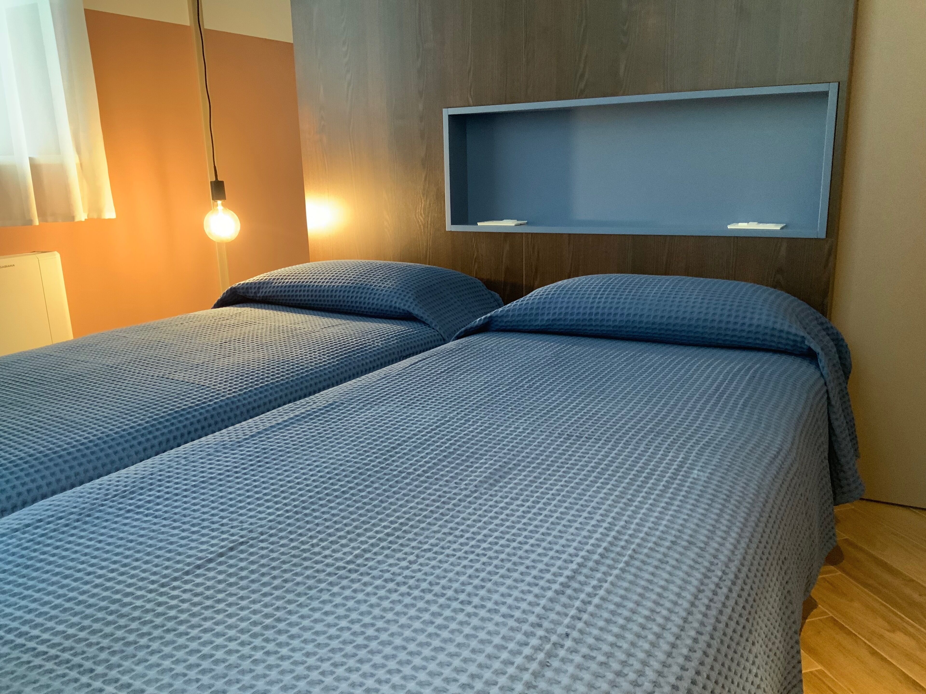 Een tweepersoonsbed met een blauwe, geruite bedspreider, een houten hoofdbord en een ingebouwd plankje erboven.