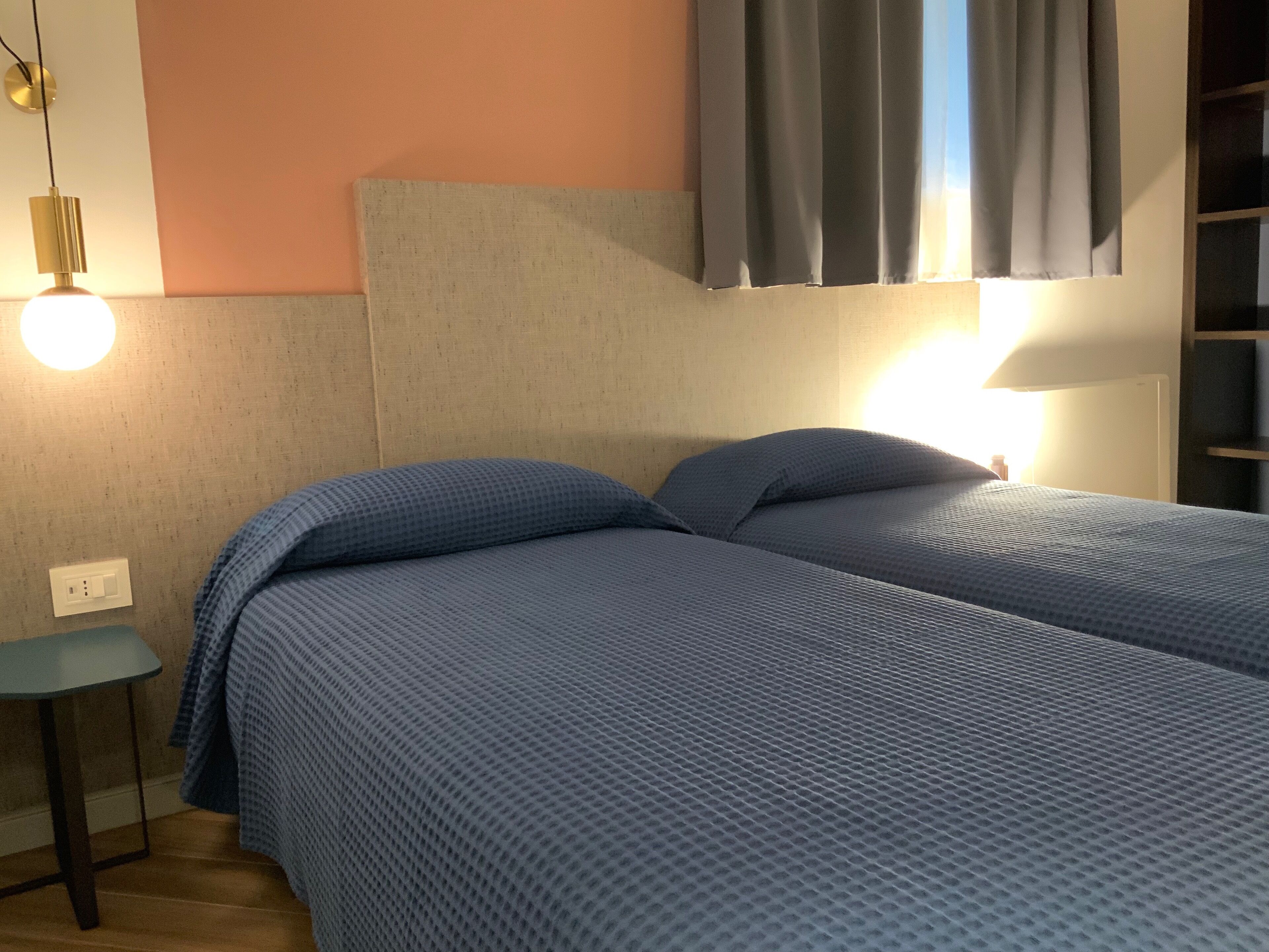 Ένα διπλό κρεβάτι με ένα μπλε κλινοσκεπάσμα με ανάγλυφη υφή, ένα κομοδίνο, ένα φωτιστικό τοποθετημένο στον τοίχο και ένα παράθυρο με κουρτίνες.