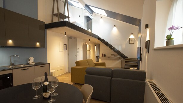A modern kitchen with a built-in oven, a dining area with a round table and wine glasses, a staircase leading to an upper level, and a living area with a sofa and a chair. - Residenza Alto Garda (Riva del Garda)