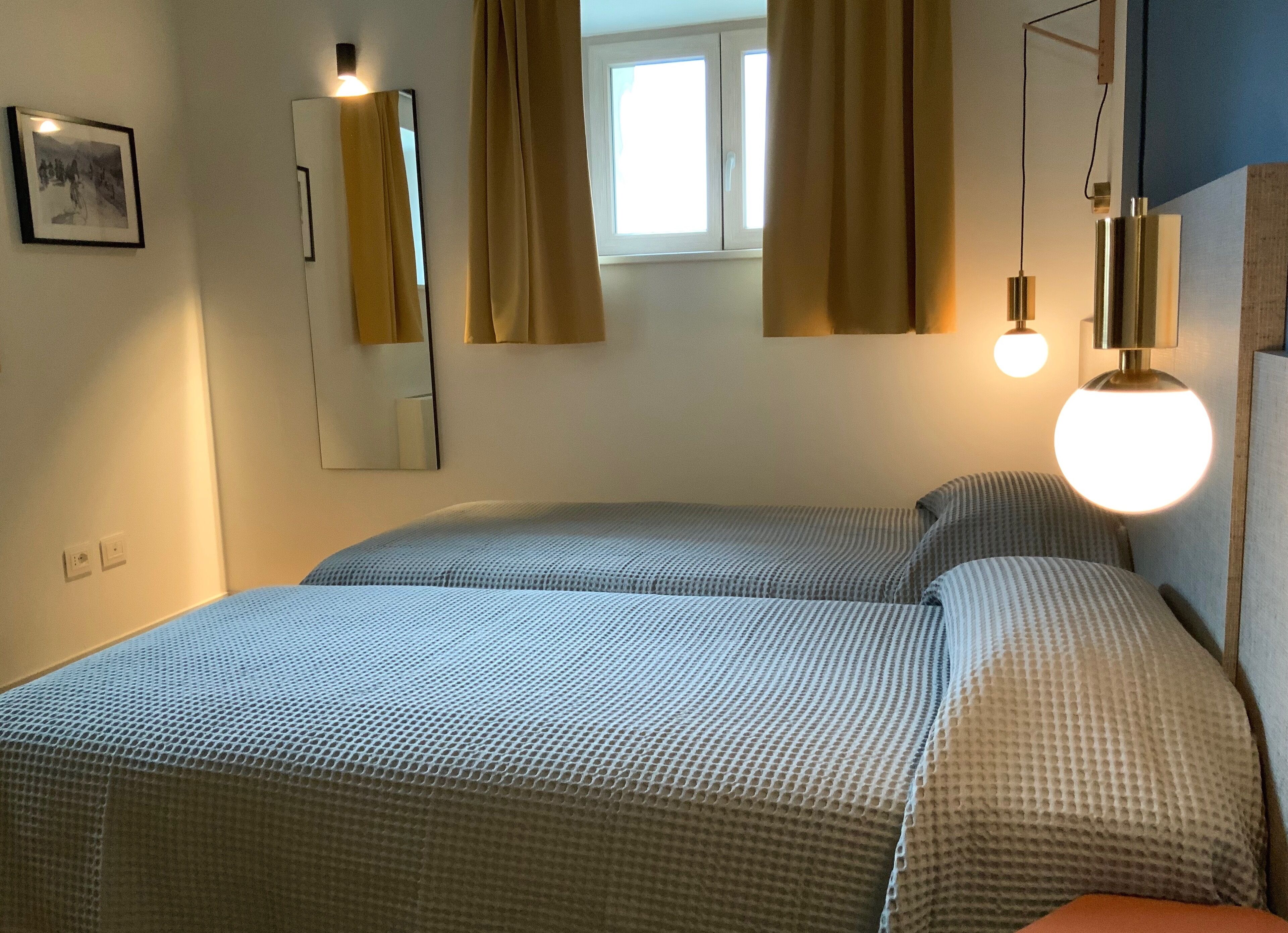 A bedroom with two beds, a mirror, a window with curtains, and a wall-mounted lamp.