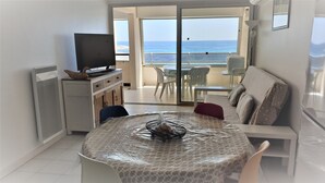 A room with a TV on a cabinet, a dining table with a plastic cover, a sofa, and a view of the sea through a glass door. - T3 in the heart of the city, on the waterfront, QUIET, Owners nearby (Le Lavandou)