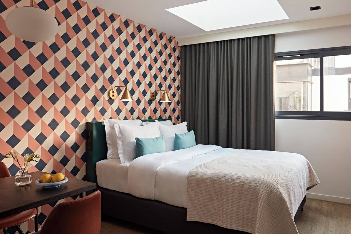 Une chambre d’hôtel moderne avec un mur orné d’un motif géométrique, un grand lit avec une literie blanche, une tête de lit sombre et une vue sur la ville par la fenêtre.