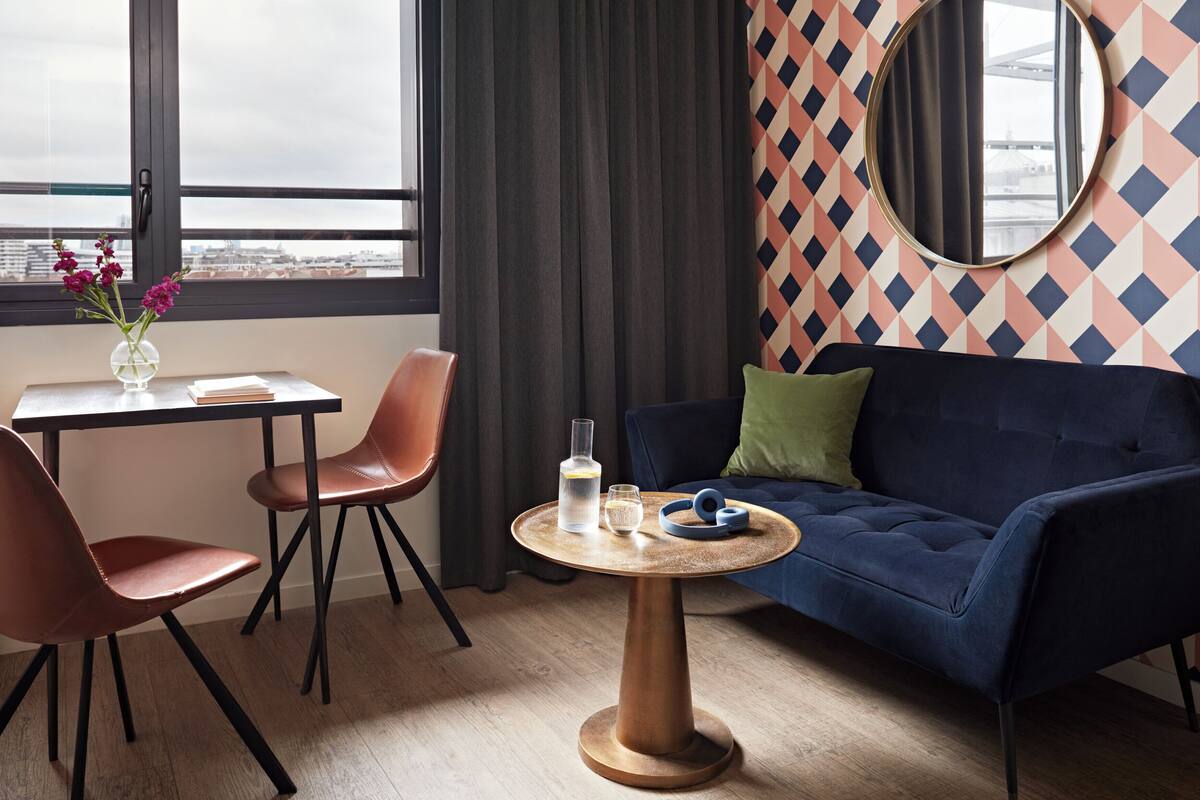 Une pièce moderne avec un canapé bleu foncé, une table ronde en bois et un petit bureau avec une chaise. Un grand miroir est accroché au mur, et une fenêtre offre une vue sur les bâtiments.