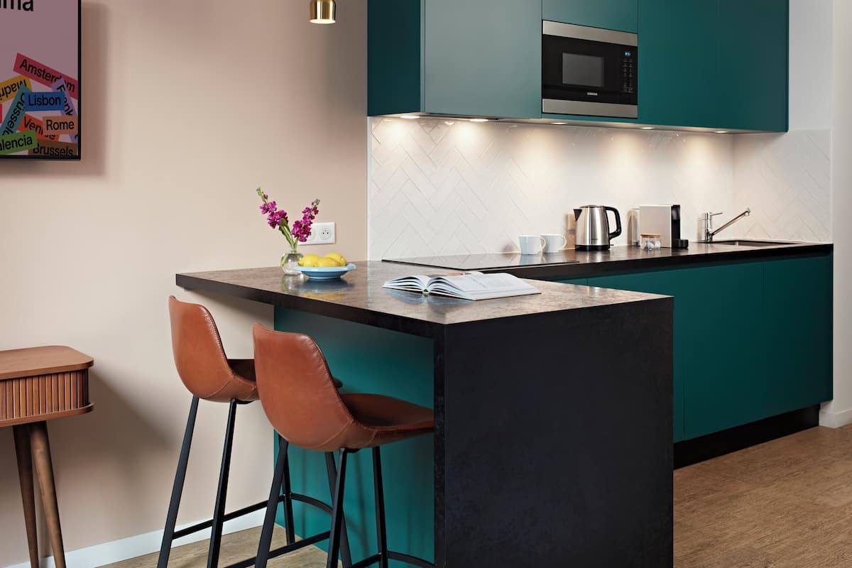 Une cuisine moderne avec des meubles de couleur turquoise, un plan de travail noir et un four à micro-ondes en acier inoxydable. On y trouve deux tabourets de bar, un vase avec des fleurs et une bouilloire sur le plan de travail.