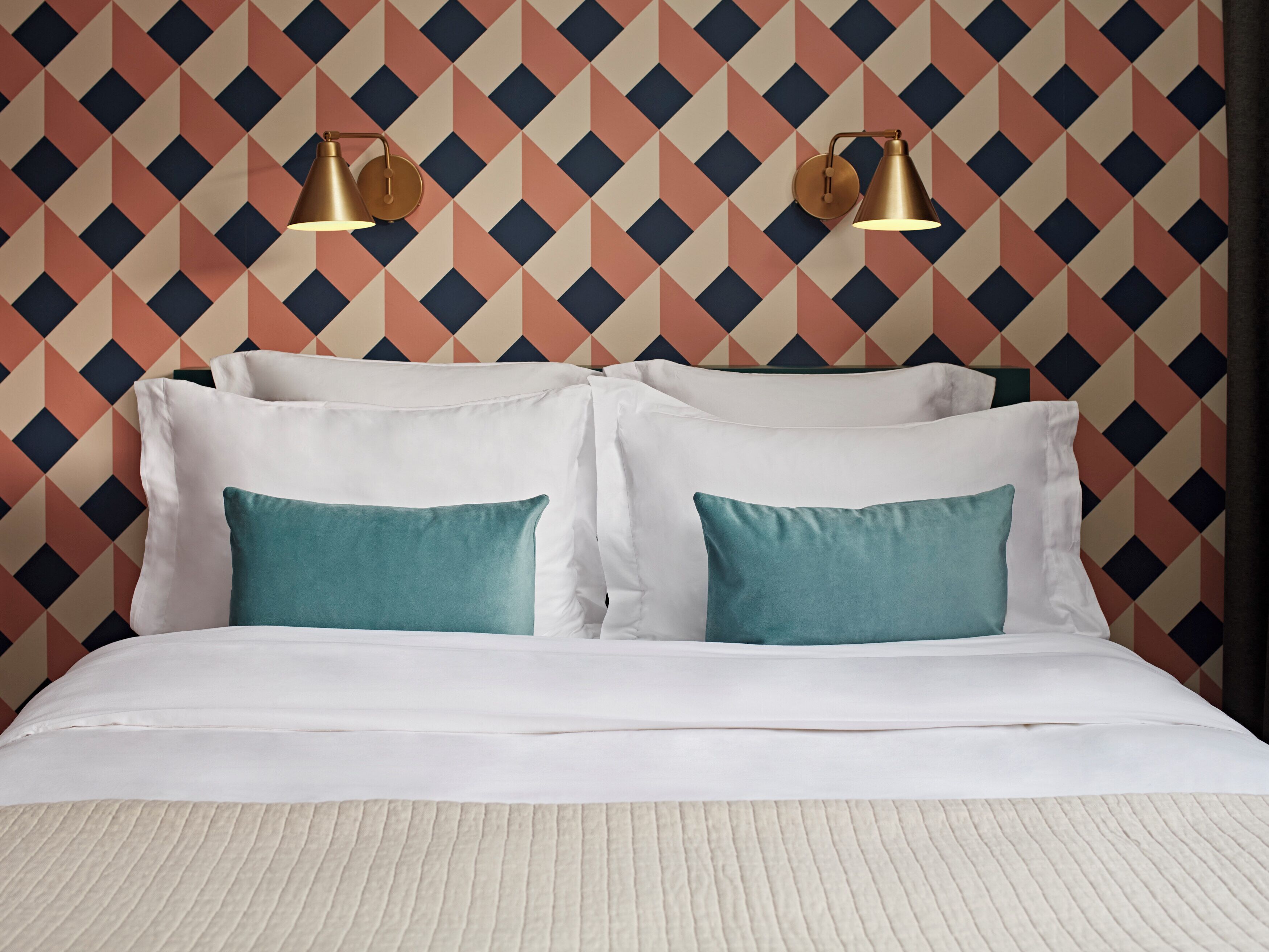 A bed with white bedding and teal pillows, set against a geometric patterned wallpaper.