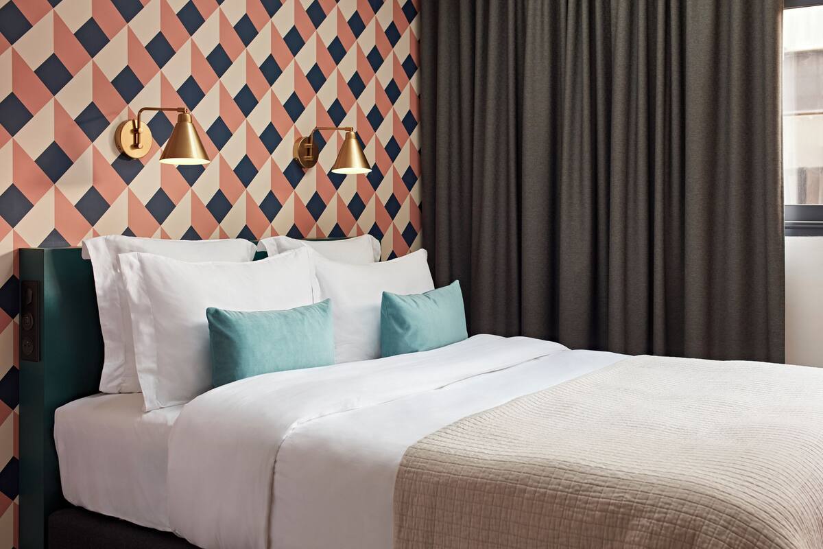 Un lit bien fait, agrémenté d’oreillers blancs et turquoise, d’une tête de lit verte et de deux lampes fixées au mur.