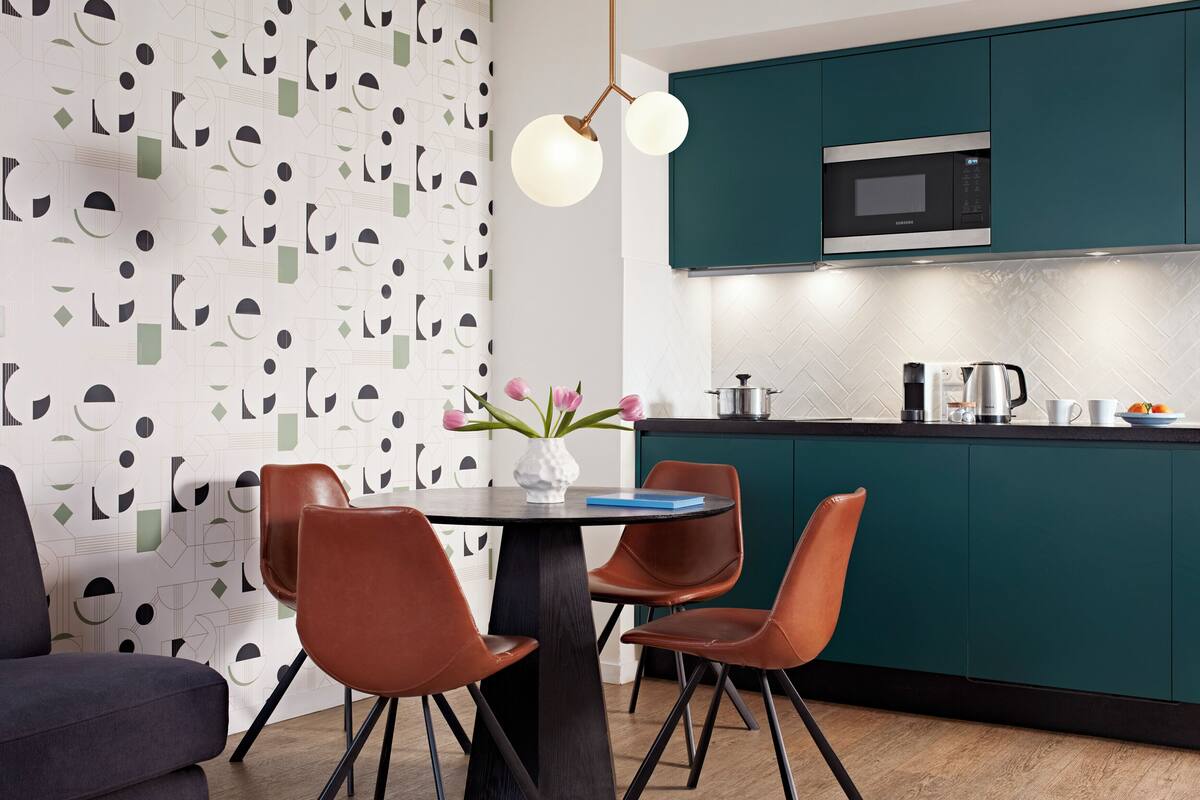 Une cuisine moderne avec des armoires de couleur turquoise, un comptoir noir et un four à micro-ondes en acier inoxydable. On y trouve une table à manger avec deux chaises marron et un vase rempli de fleurs.