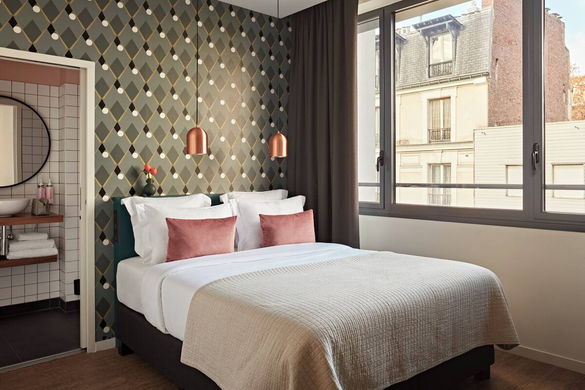 Une chambre d’hôtel moderne dotée d’un grand lit, d’une salle de bain avec un miroir et un lavabo, et offrant une vue sur la ville par une fenêtre.