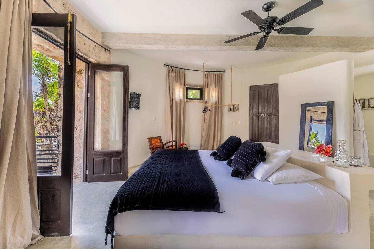 A bedroom with a large bed, a ceiling fan, a window with curtains, and a view of greenery outside.
