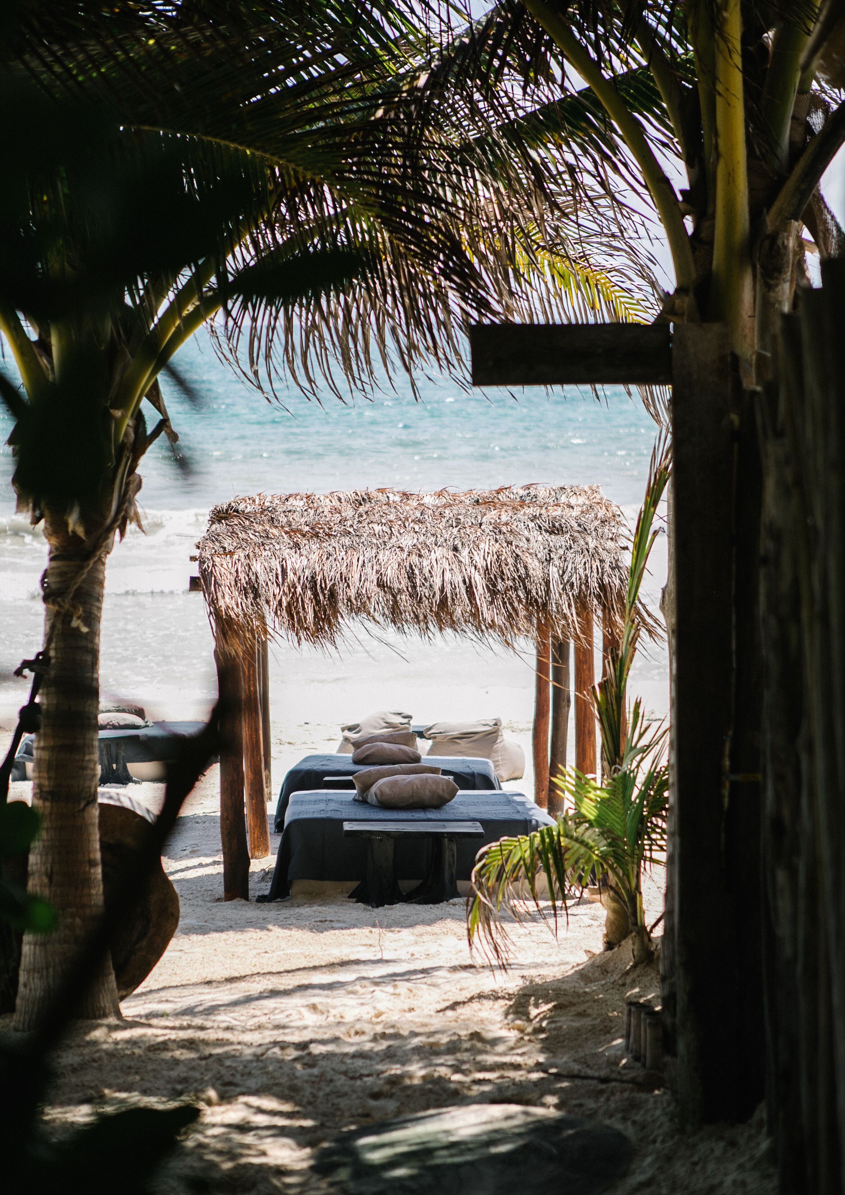 A beachside hut with a thatched roof, a table with cushions, and a view of the ocean.