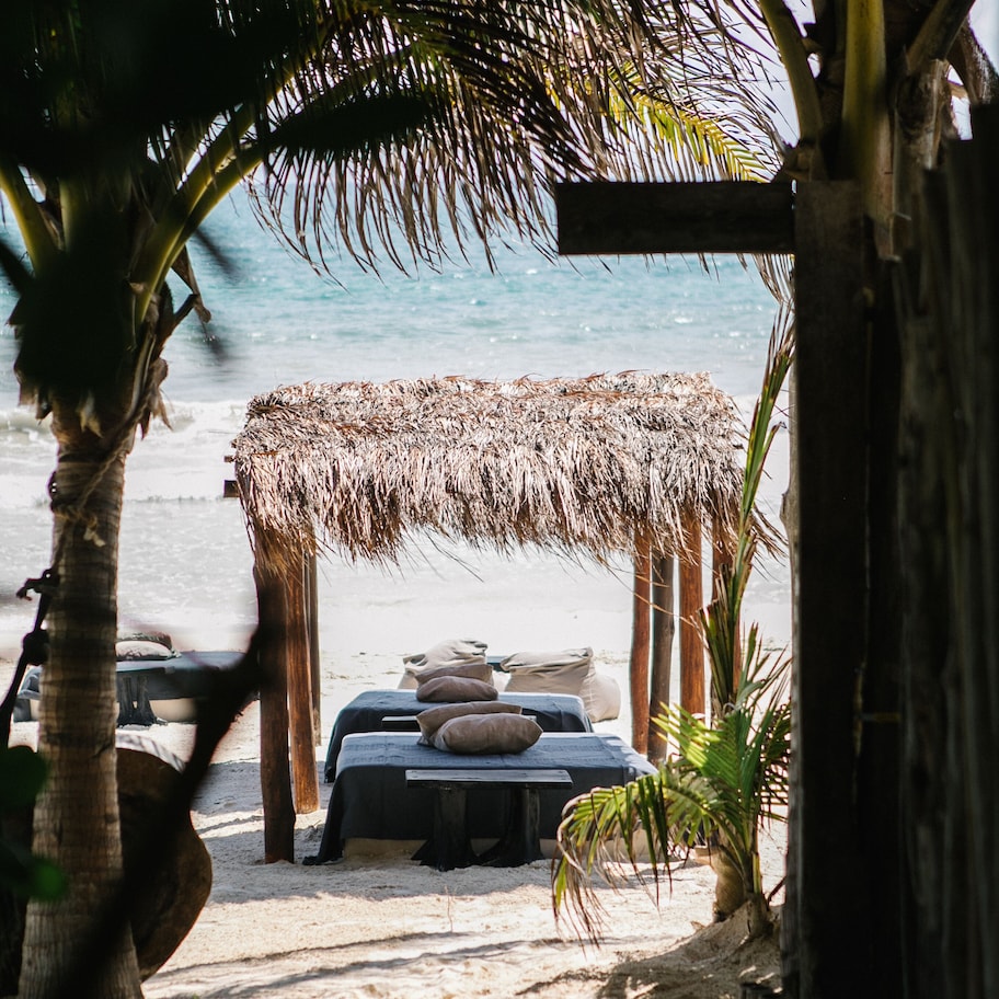 A beachside hut with a thatched roof, a table with cushions, and a view of the ocean.