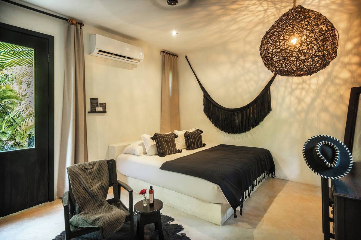 A bedroom with a bed, a chair, a small table with a bottle and flowers, an air conditioning unit, a hanging light fixture, and a window with curtains.
