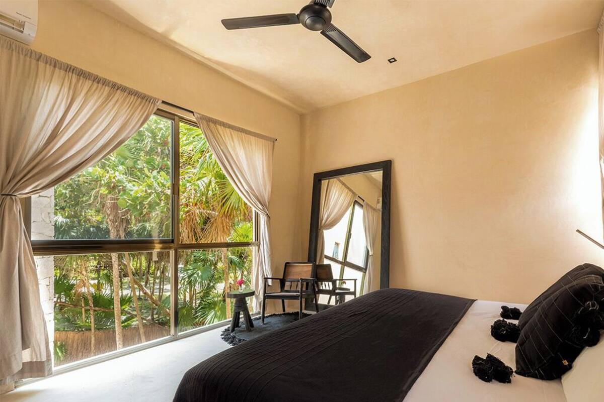 A bedroom with a large bed, a ceiling fan, a window with curtains, and a view of greenery outside.