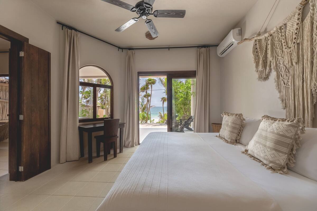 A bedroom with a large bed, a ceiling fan, an air conditioning unit, a desk with a chair, and a view of the beach and palm trees through a glass door.