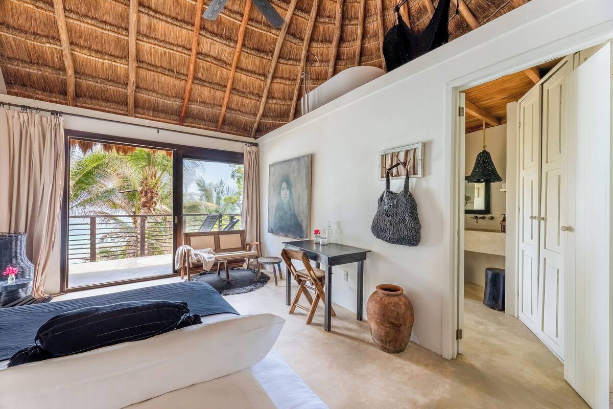 A bedroom with a bed, a desk, a chair, a large vase, and a view of palm trees through the window.